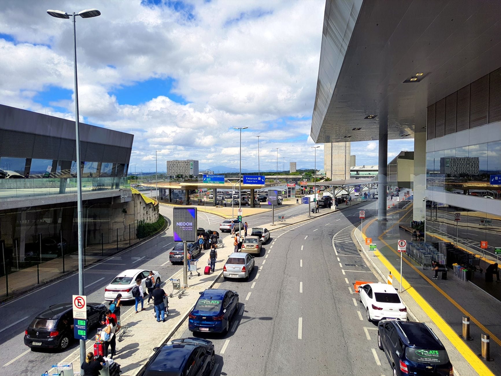 Área de desembarque movimentada com carros, táxis, passageiros e o terminal ao fundo sob um céu claro em um aeroporto internacional.