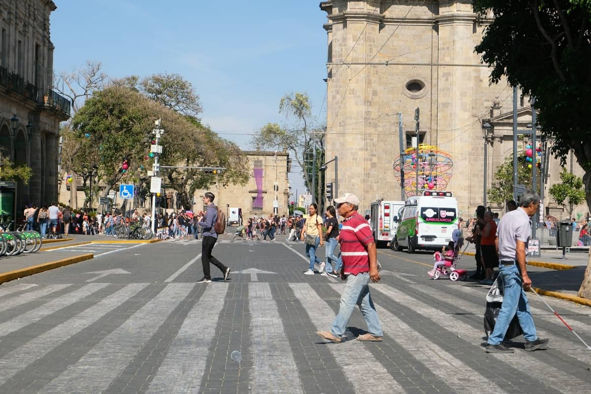 Menschen überqueren eine belebte Innenstadtstraße nahe historischer Steingebäude im Zentrum von Guadalajara an einem sonnigen Tag.