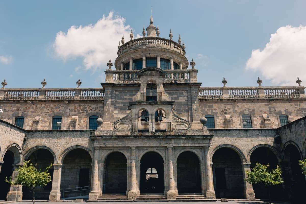 Vorderansicht des Hospicio Cabañas in Guadalajara, mit seiner beeindruckenden Architektur und der zentralen Kuppel unter einem blauen Himmel mit Wolken.