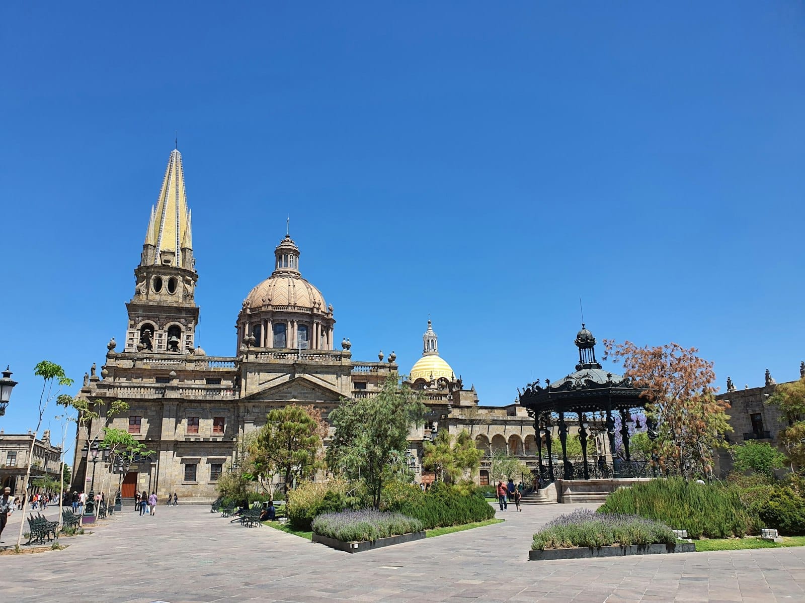 Vista ampla da Catedral de Guadalajara com suas icônicas torres e cúpulas amarelas, um coreto em primeiro plano e pessoas caminhando numa praça ensolarada.