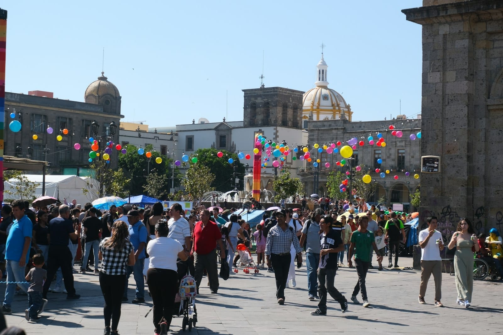 Multidão caminha por uma praça festiva no centro de Guadalajara, com bandeirolas coloridas de papel picado penduradas e as cúpulas da catedral histórica ao fundo.