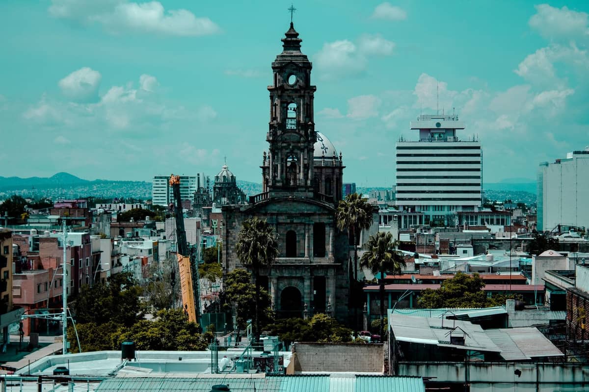 Luftaufnahme der Stadtlandschaft von Guadalajara mit einem historischen Kirchturm, umgeben von städtischen Gebäuden unter einem bewölkten blauen Himmel.