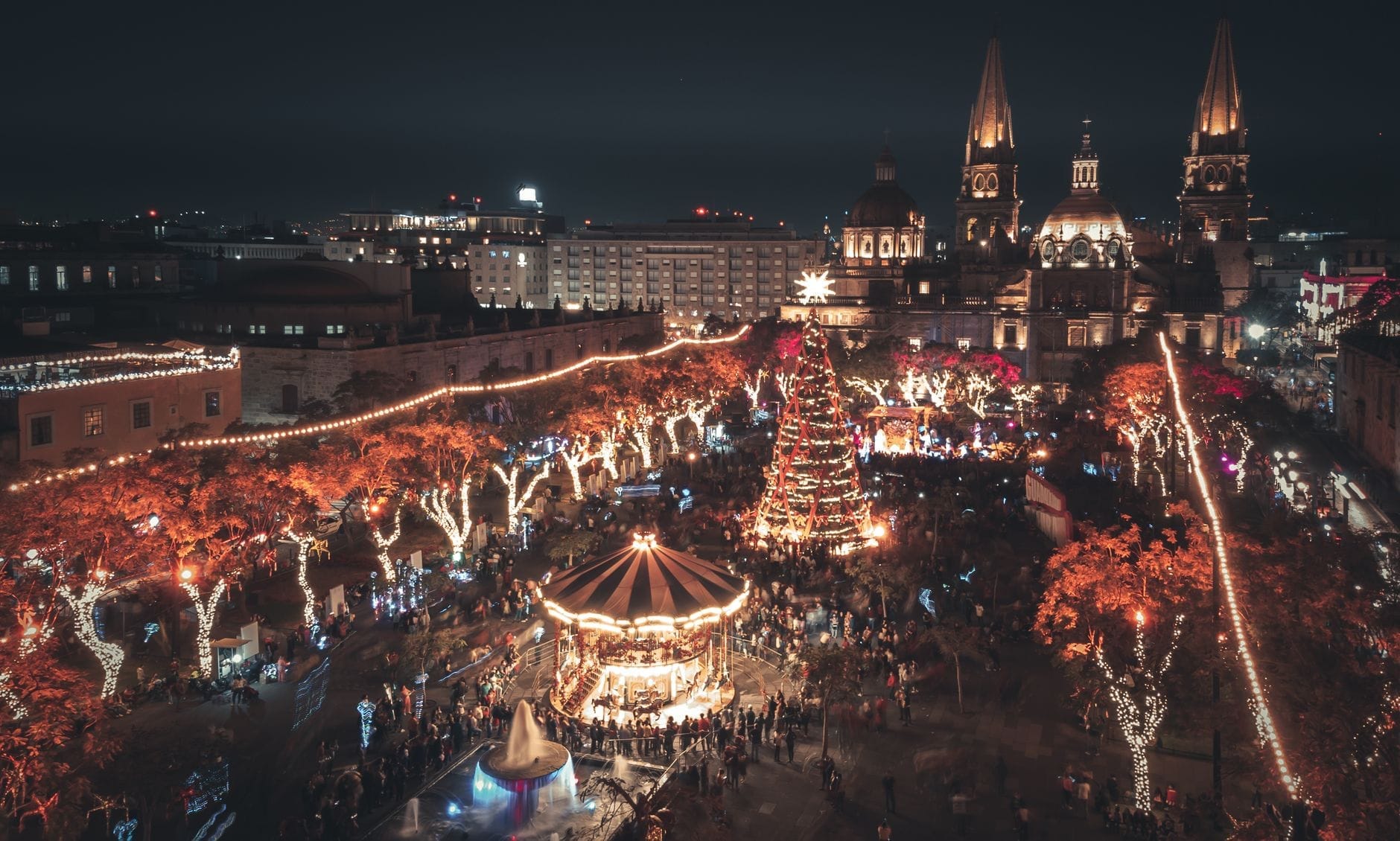 Vista aérea da praça principal de Guadalajara à noite, iluminada com luzes festivas, uma grande árvore de Natal, carrossel e multidões reunidas perto da catedral.