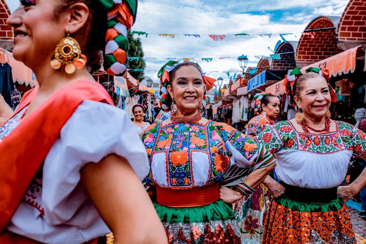 Frauen in traditionellen mexikanischen Kleidern, die lächeln und auf einem festlichen Freiluftmarkt mit bunten Dekorationen tanzen.