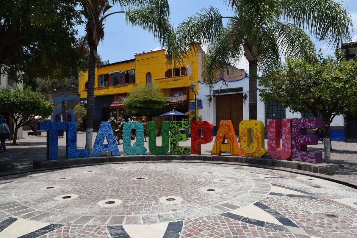 Buntes Tlaquepaque-Schild vor Palmen und lebhaften Gebäuden auf einem belebten Platz in Tlaquepaque, Guadalajara.