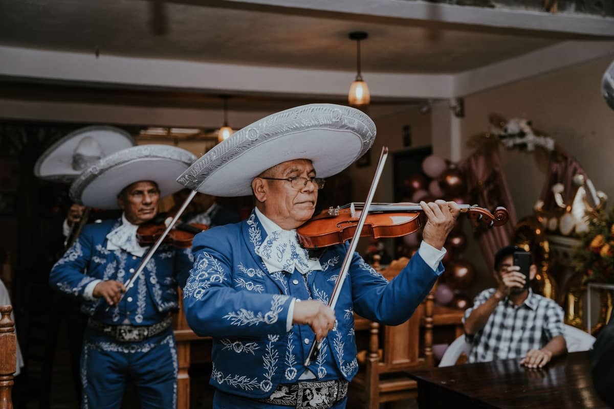 Mariachi-Musiker in traditionellen blauen Anzügen spielen Geigen in einem dekorierten Raum mit sitzenden Gästen und festlicher Beleuchtung.
