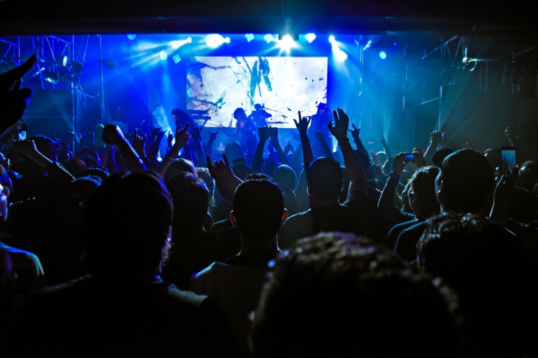 Multidão dançando e levantando as mãos em uma balada animada com luzes azuis no palco e atmosfera cheia de energia à noite.