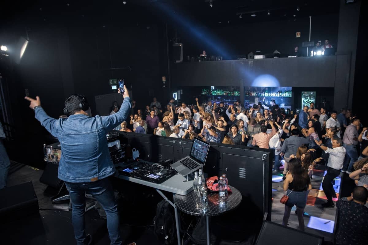 DJ legt für eine lebhafte Menge auf einer Tanzfläche in einem modernen Club auf, während Menschen tanzen und sich unterhalten.