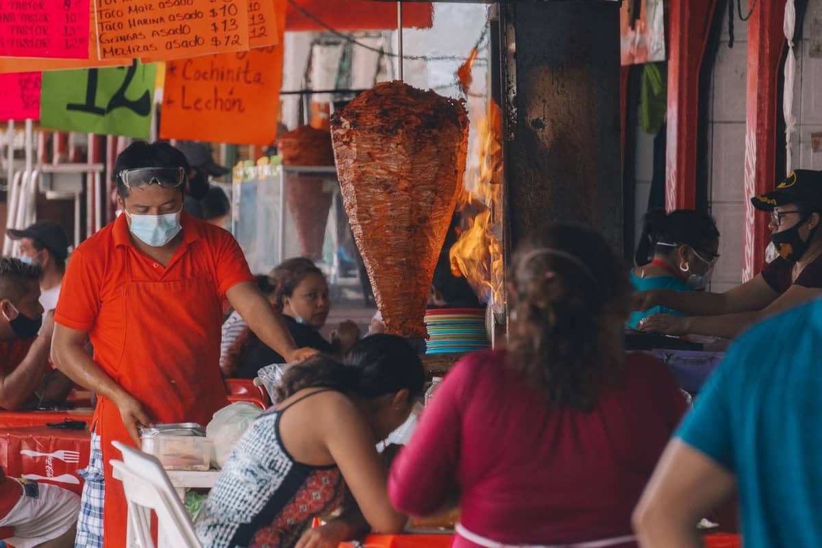 Belebter Straßenimbissstand in Guadalajara mit essenden Menschen, einem servierenden Koch und einem großen Trompo mit über Flammen geröstetem Fleisch.
