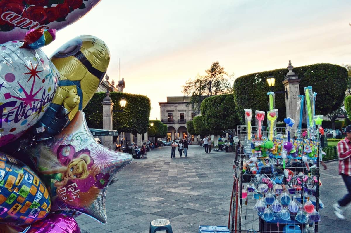 Ein lebhafter Platz in Guadalajara bei Sonnenuntergang, mit Luftballons und Straßenhändlern im Vordergrund und Menschen, die sich in der Nähe gestutzter Bäume versammeln.