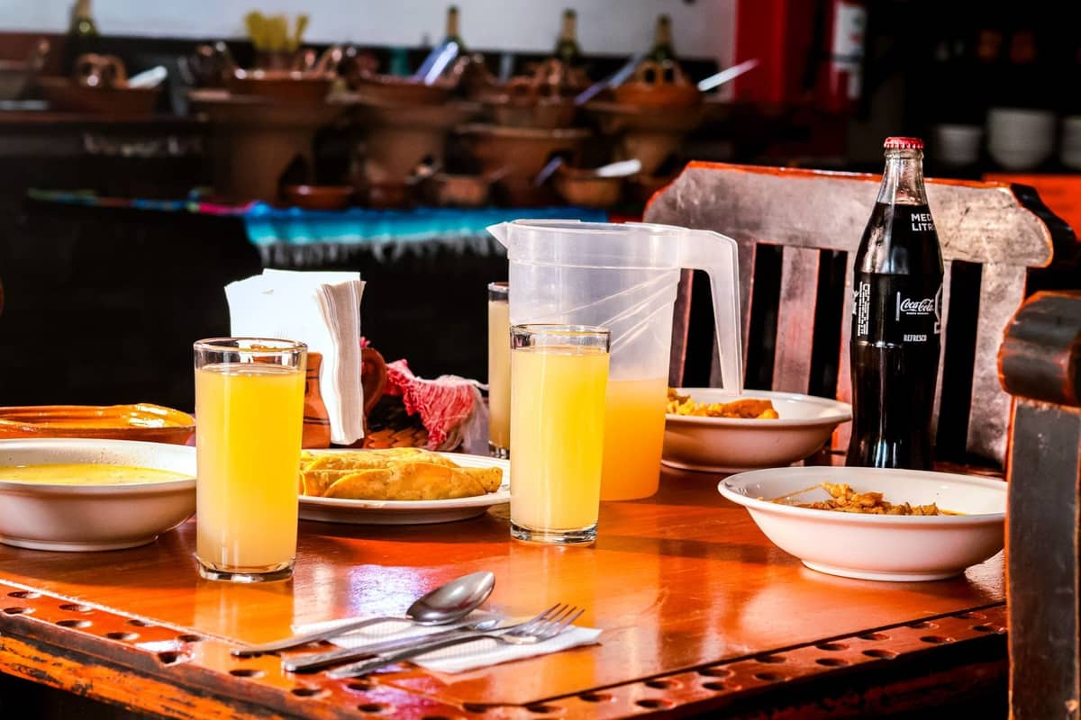 Ein rustikaler Restauranttisch mit Schüsseln Eintopf, Gläsern Saft und einer Glasflasche Limonade, der das lokale Speiseerlebnis in Mexiko widerspiegelt.