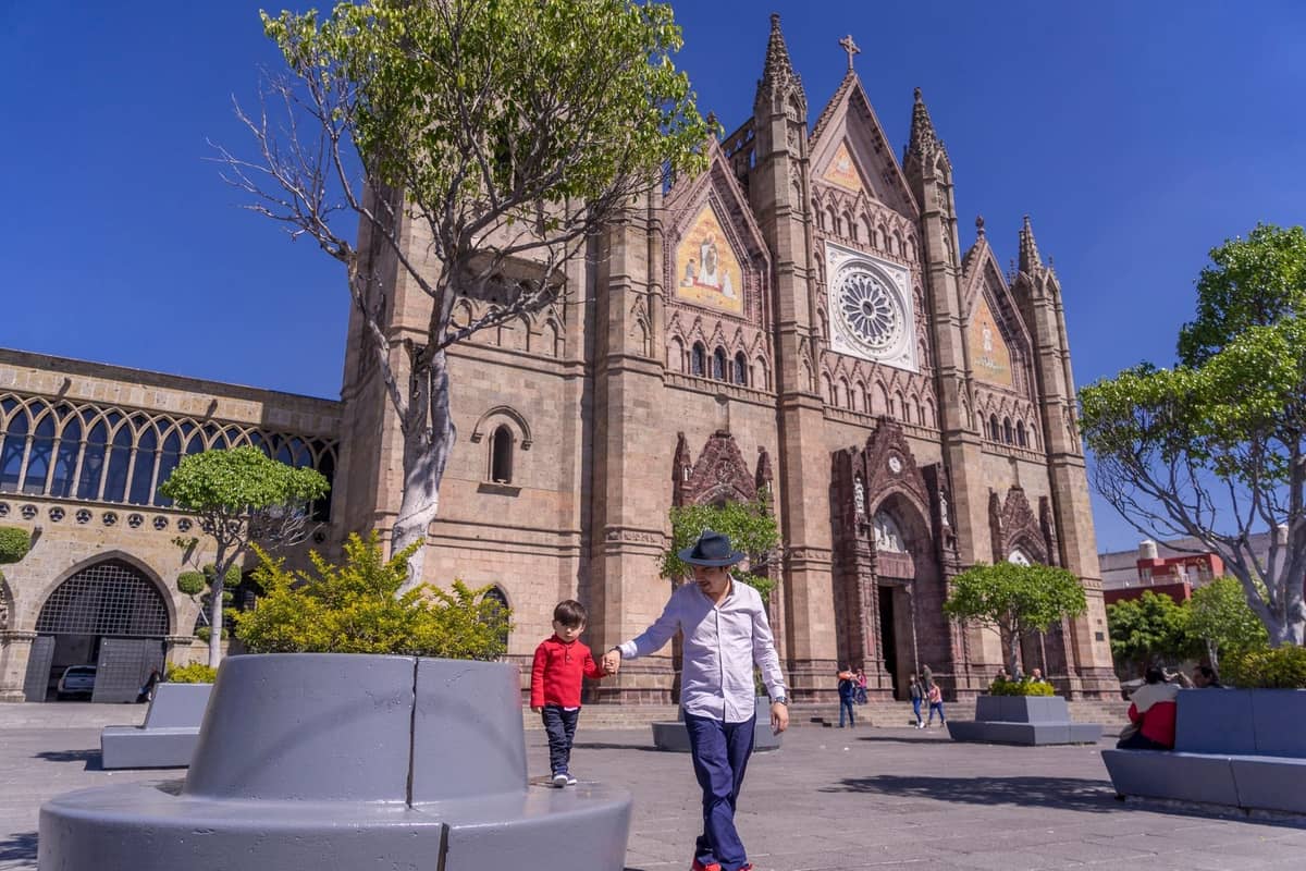 Kinder spielen auf einem sonnigen Plaza vor dem Templo Expiatorio, einer bekannten gotischen Kirche in Guadalajara, während Familien die Zeit im Freien genießen.