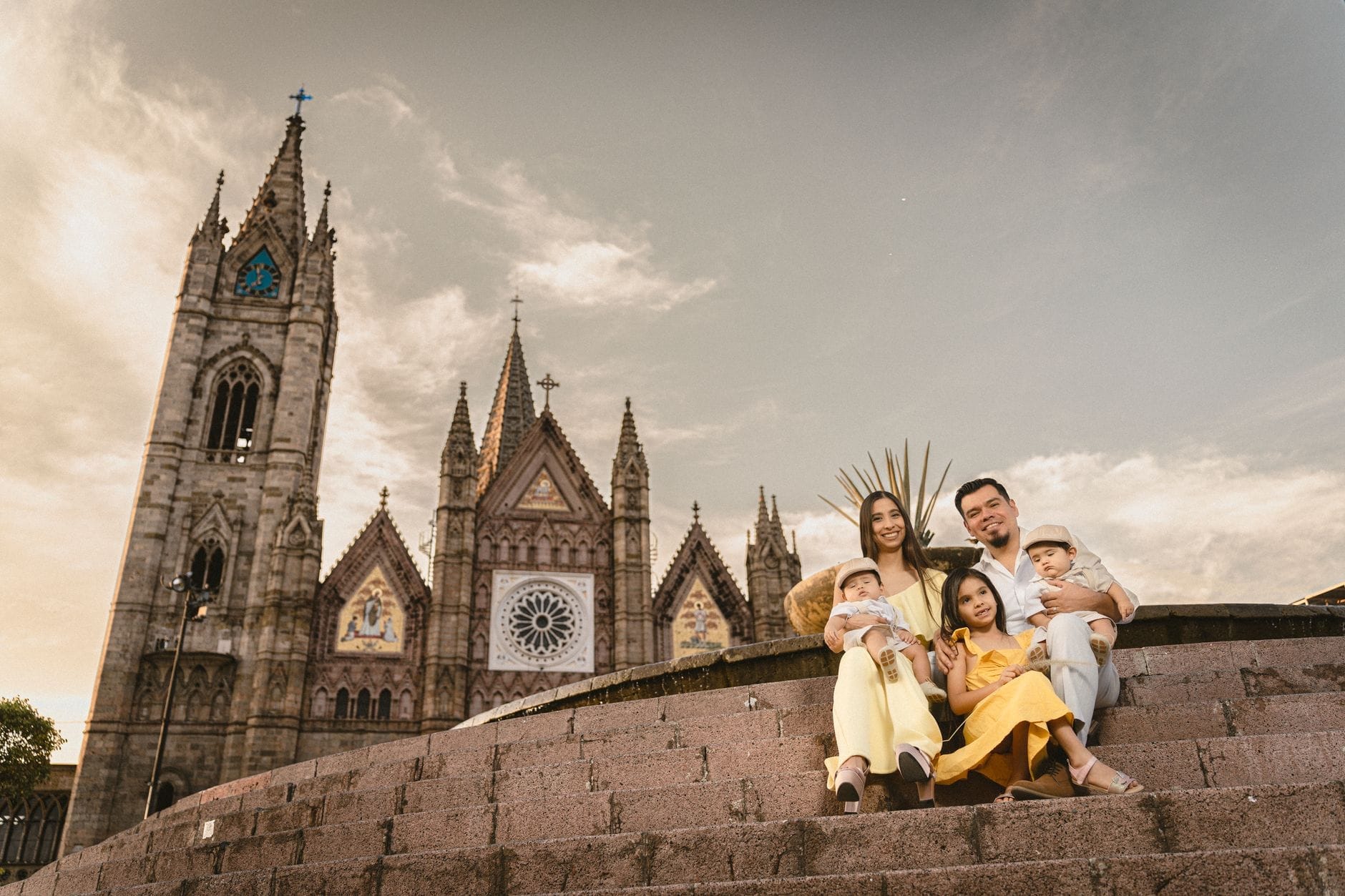 Família sorridente com três filhos pequenos sentada nos degraus de pedra em frente ao icônico Templo Expiatorio neogótico de Guadalajara, sob um céu dramático ao entardecer.