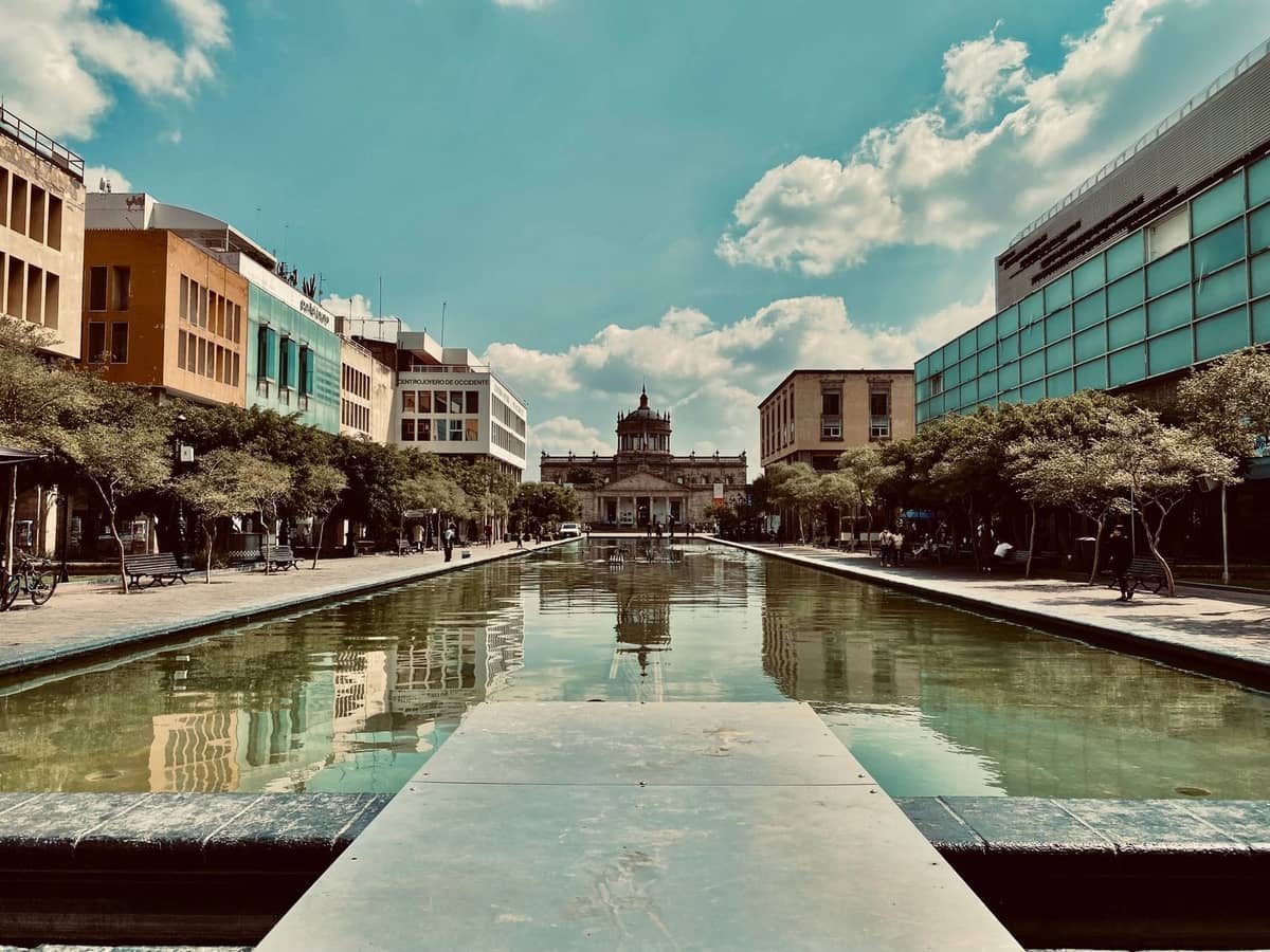 Blick über einen breiten Platz mit einem Spiegelbecken, der zum historischen Hospicio Cabañas in der Innenstadt von Guadalajara unter blauem Himmel führt.
