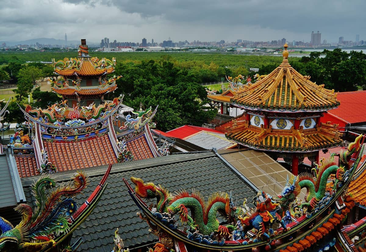 Verzierte, mit Drachen geschmückte Dächer des Guandu-Tempels in Taipei, mit üppigen grünen Feuchtgebieten und der Skyline der Stadt unter einem bewölkten Himmel.