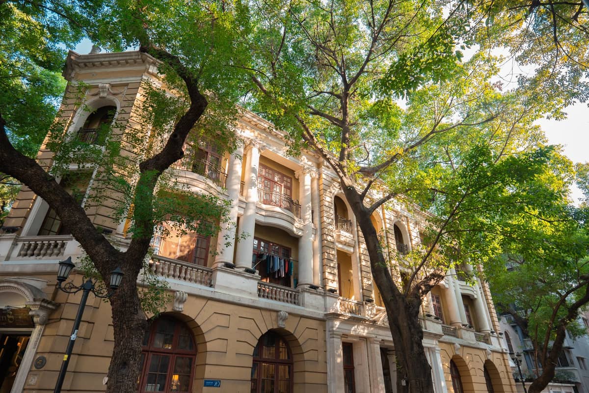 Edifício histórico de estilo colonial na Ilha Shamian em Guangzhou, com características neoclássicas, janelas em arco e rua pedonal arborizada em primeiro plano.