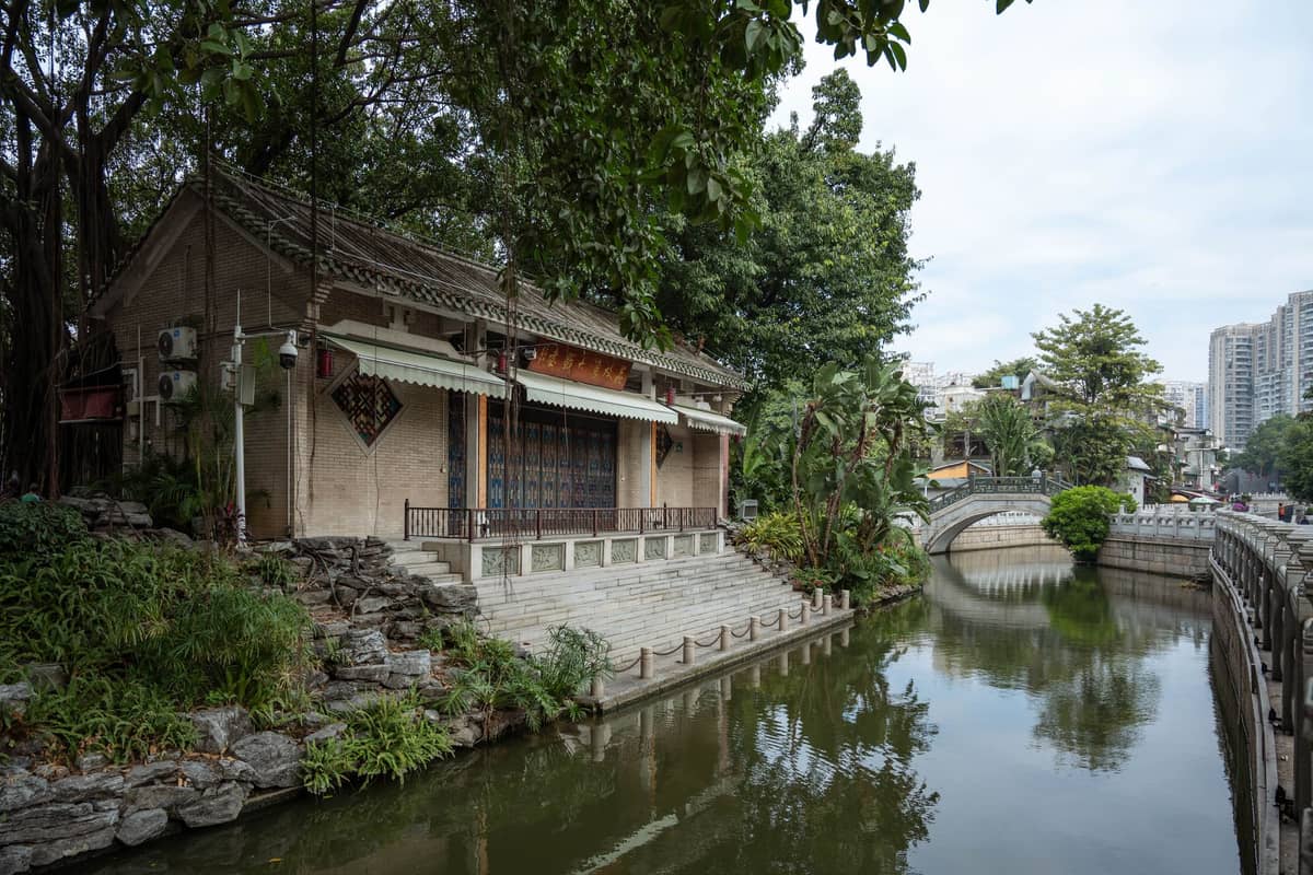 Edifício tradicional de estilo Lingnan com telhado de telhas e varanda aberta ao lado de um canal sombreado por árvores em Guangzhou.