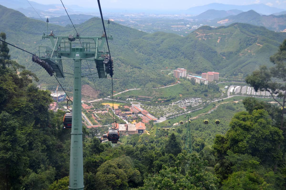Teleféricos subindo uma montanha verde com edifícios da cidade e colinas visíveis ao longe abaixo do elevador.