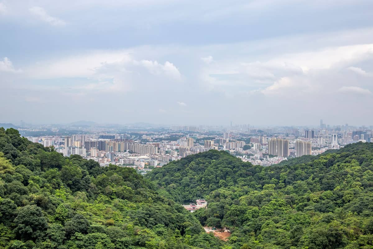 Colinas arborizadas e exuberantes de Baiyun Mountain com o horizonte urbano de Guangzhou visível ao fundo sob um céu parcialmente nublado