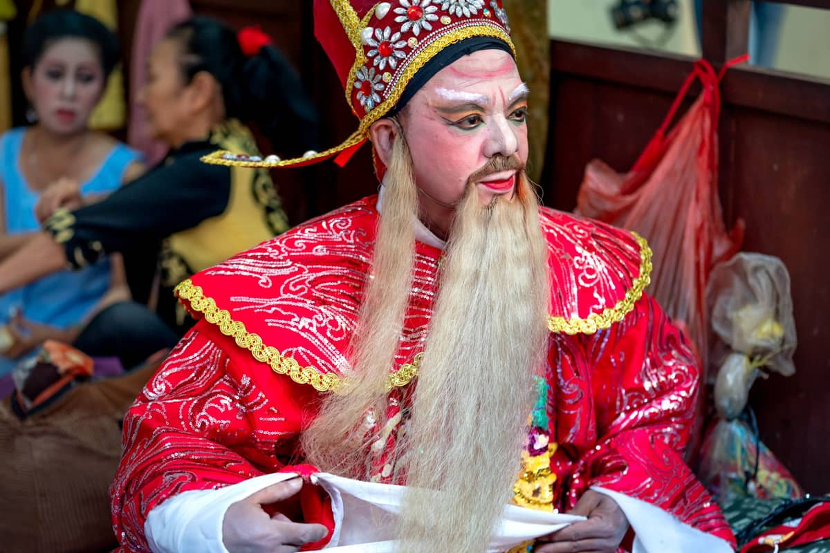 Cantonese opera performer in traditional ornate costume and dramatic face makeup, sitting backstage with other performers preparing in the background.
