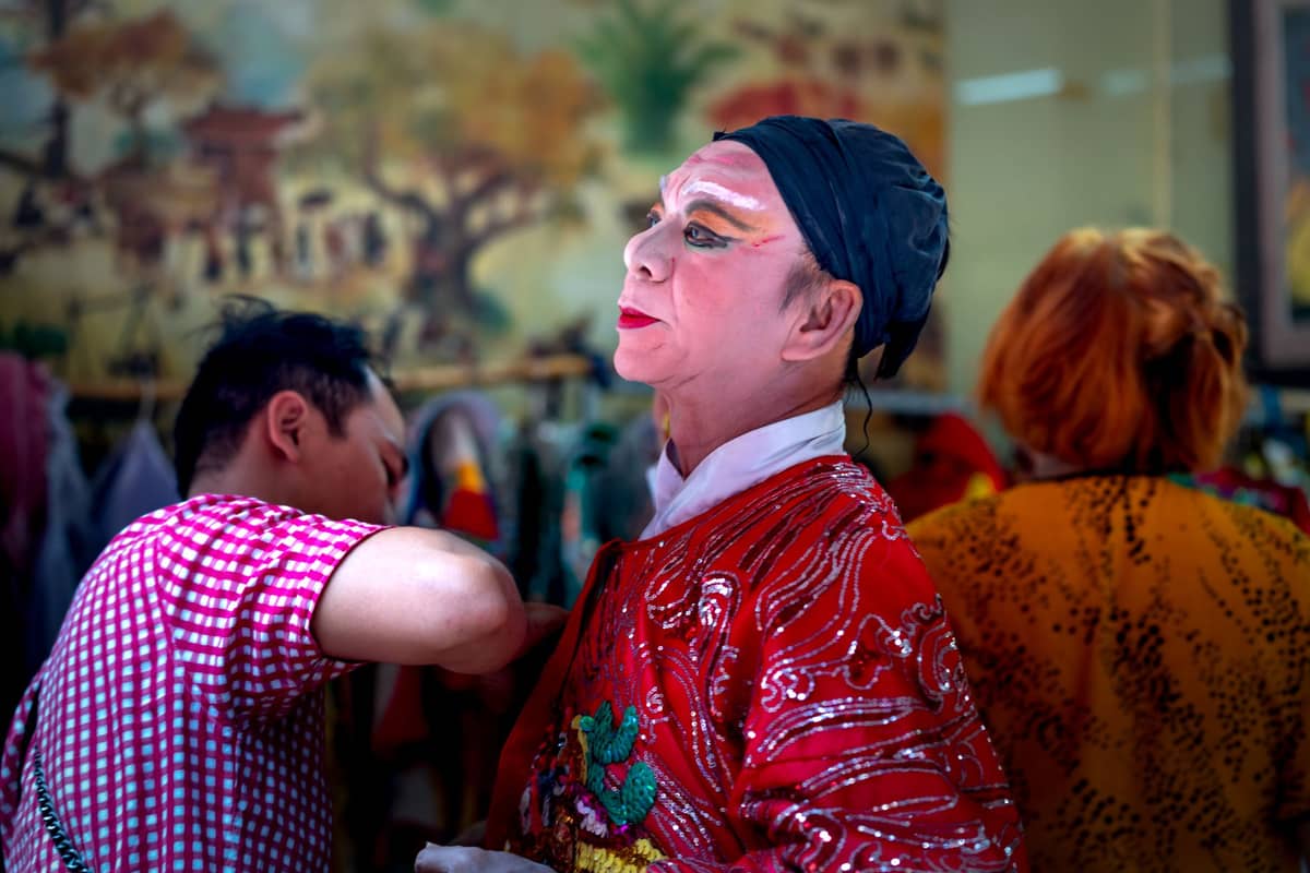 Performer in elaborate embroidered Cantonese opera costume and makeup being dressed backstage, with painted scenery in the background.