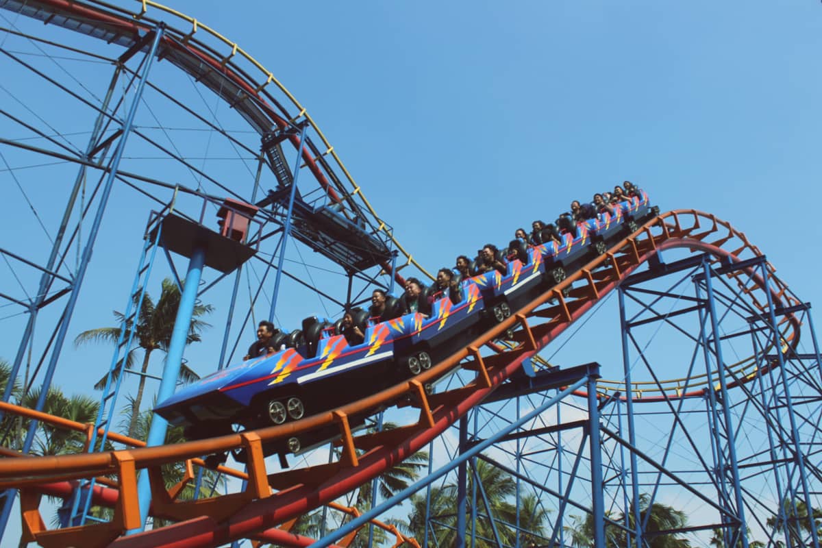 A brightly colored roller coaster with riders heads downward on a steep drop, surrounded by blue support beams and palm trees, under a clear sky.