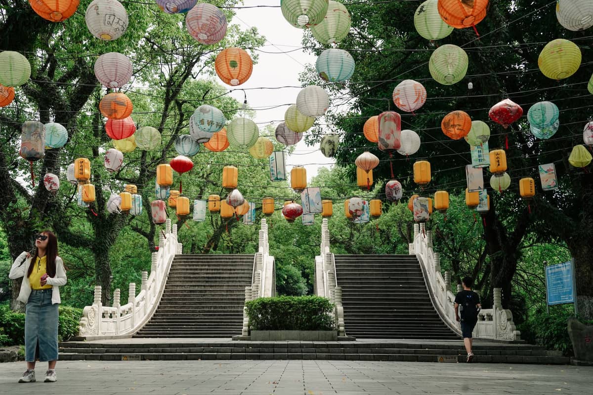Stone bridge stairs in a park with colorful lanterns overhead, people walking, lush green trees all around.