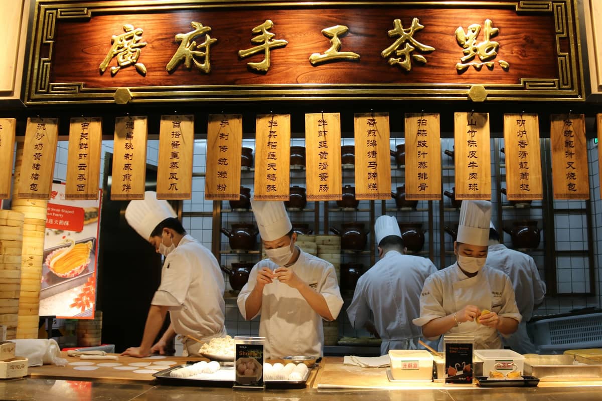 Four chefs wearing white uniforms and hats prepare dim sum behind a wooden counter under a traditional Cantonese sign.
