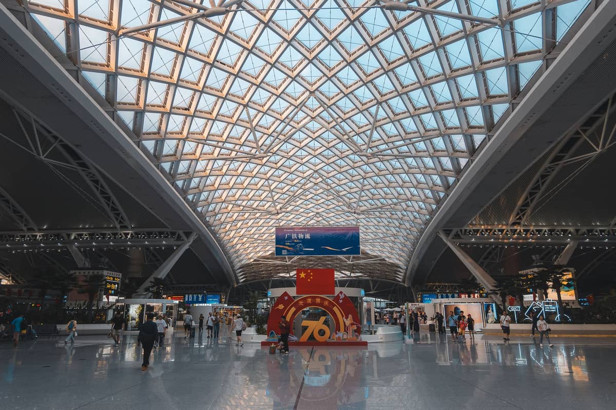 Interior of a modern airport terminal with a high glass roof, travelers, and a large decorative display in the center.