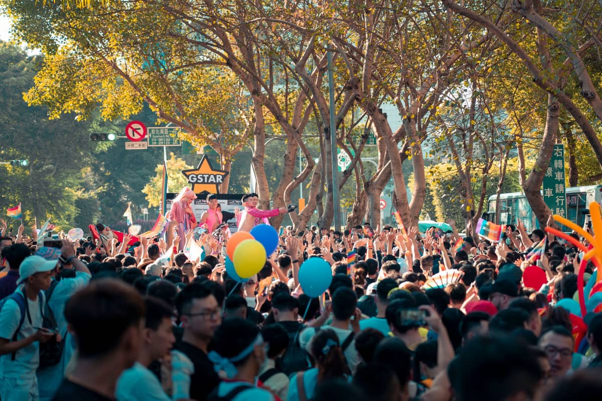 A large outdoor crowd at a festival, with balloons and performers on stilts, under trees and city street signs.