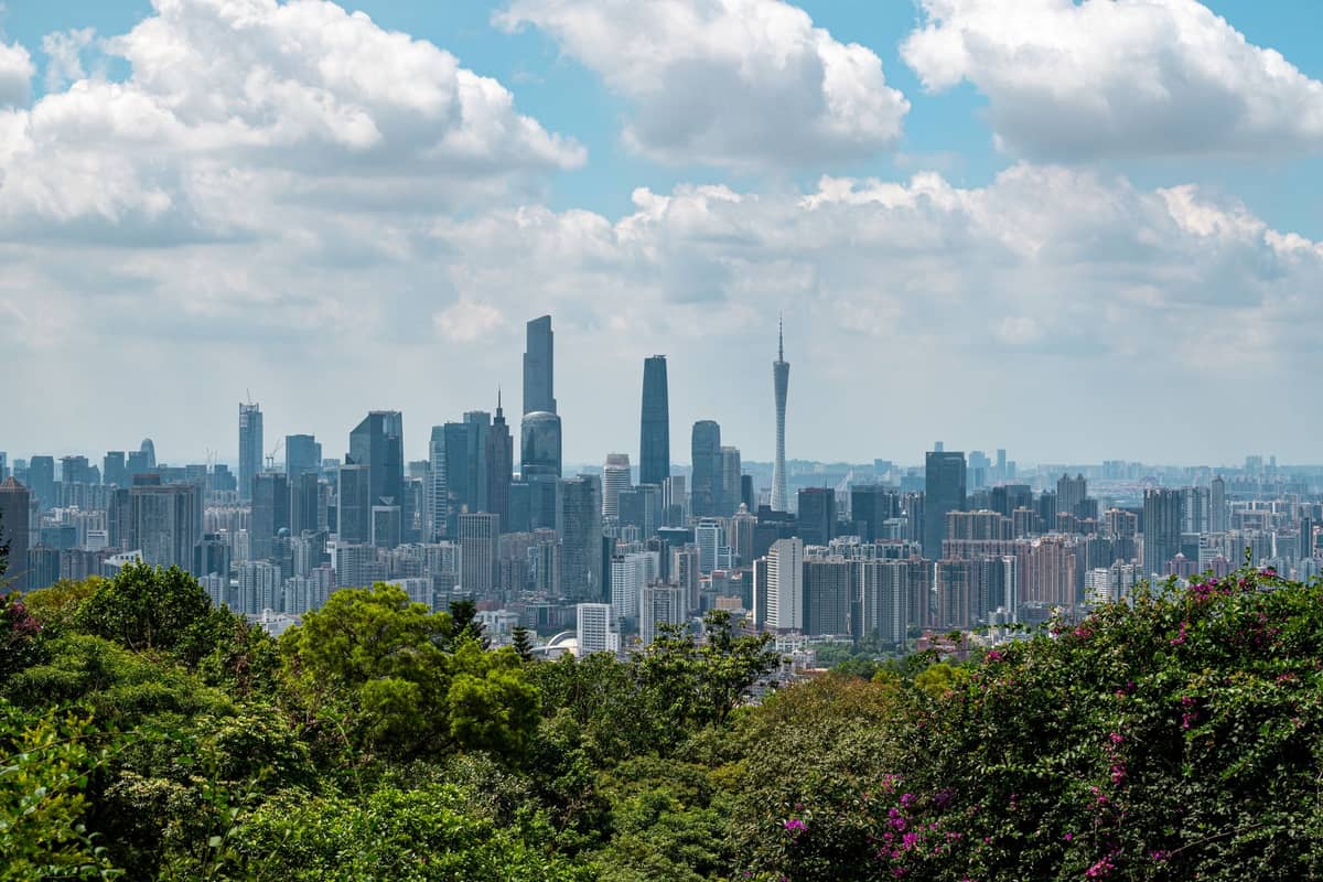 Panoramic daytime view of Guangzhou skyline with lush greenery in the foreground and modern skyscrapers, including Canton Tower, under partly cloudy skies.
