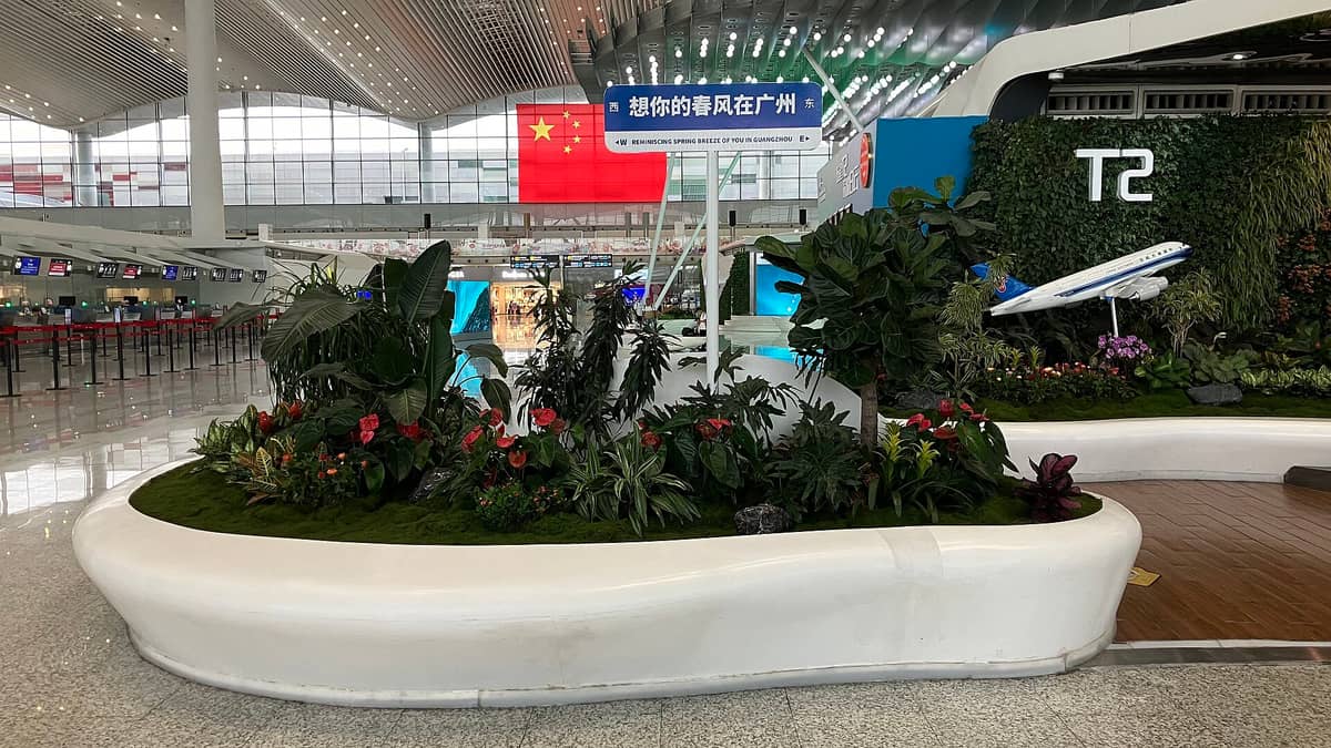Interior of Guangzhou Baiyun International Airport with modern architecture, lush indoor greenery, check-in counters, and a sign referencing Guangzhou.
