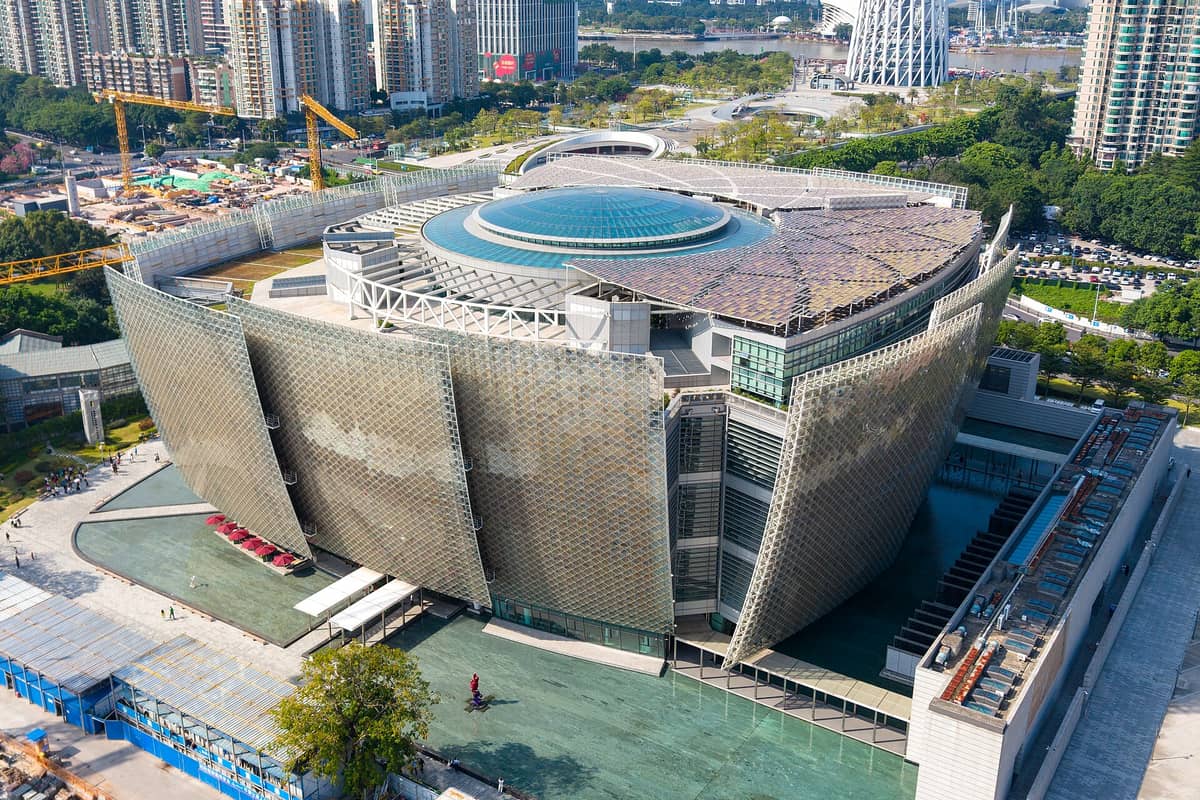 Aerial view of the Guangzhou Museum of Art’s striking modern building with distinctive glass dome and angular exterior panels, surrounded by city buildings and green trees.