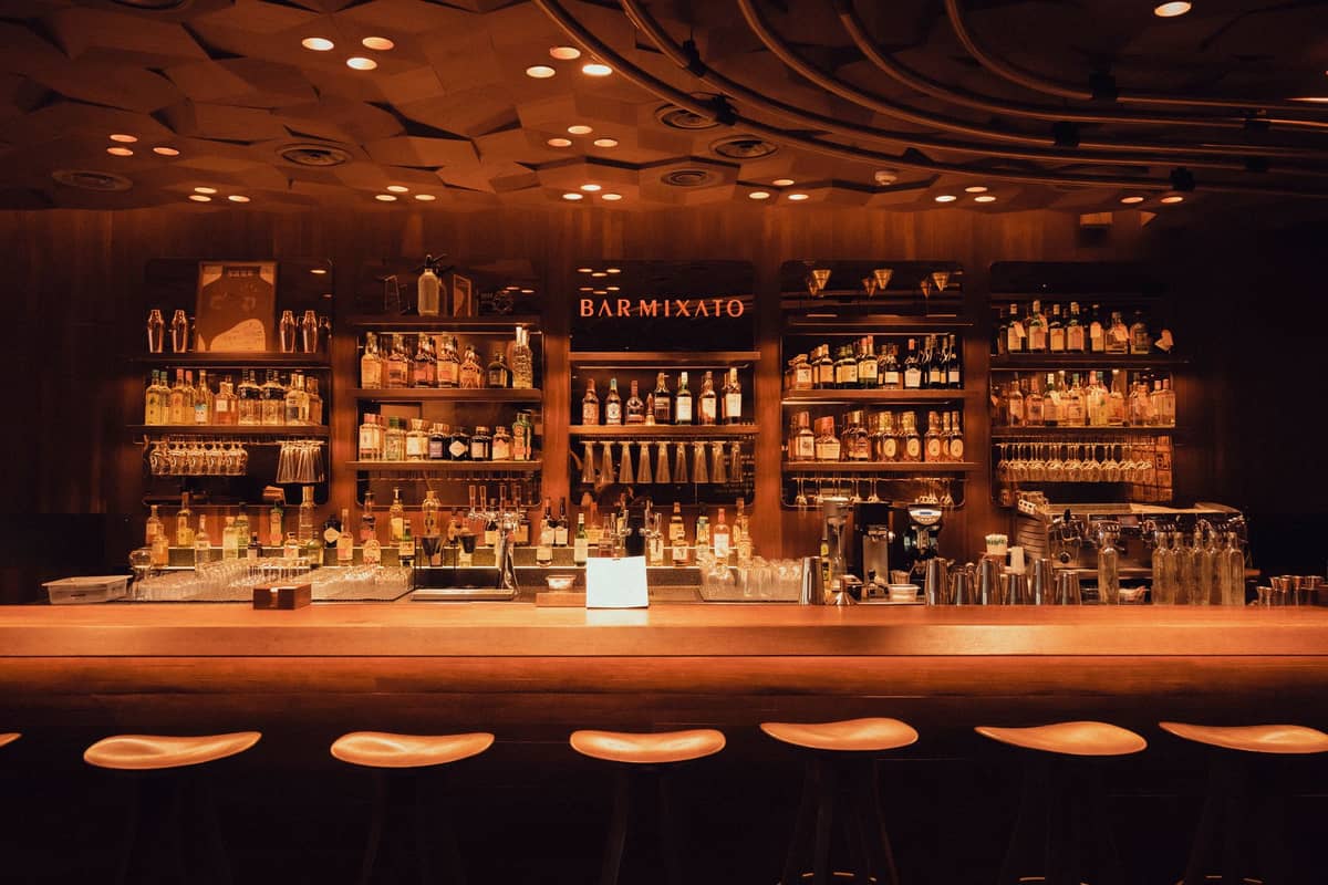 Atmospheric cocktail bar with shelves of bottles, warm lighting, and a polished bar counter with stools in front