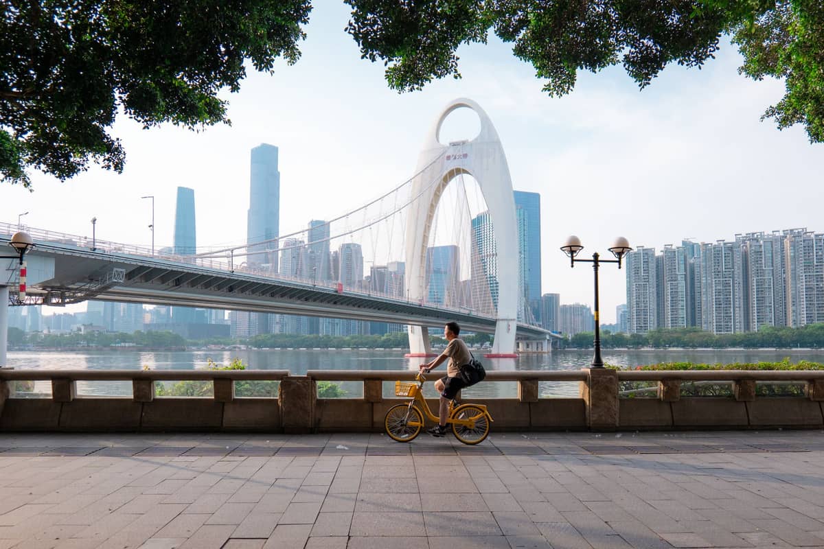 Daytime waterfront promenade in Guangzhou with the distinctive Liede Bridge, city skyline, and a cyclist on a shared bike