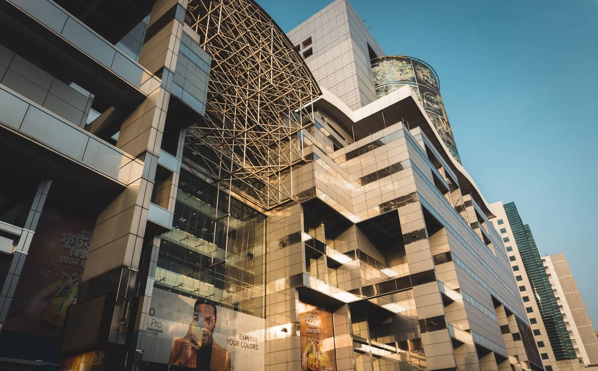 Modern shopping mall with glass and metal facade in an urban area circled by high rises under a clear sky, likely in a major city.