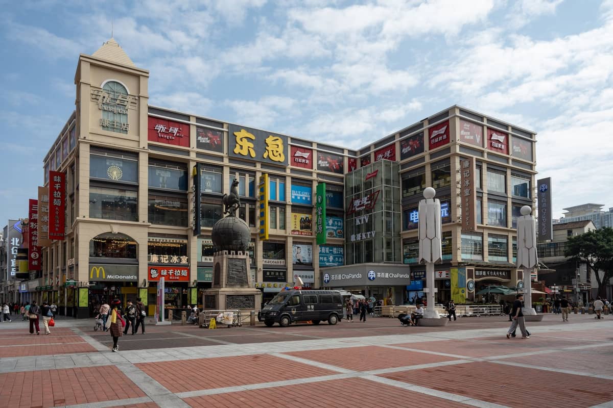Multi-story shopping mall with colorful signs and shopfronts in a large public plaza in Guangzhou, busy with people walking.
