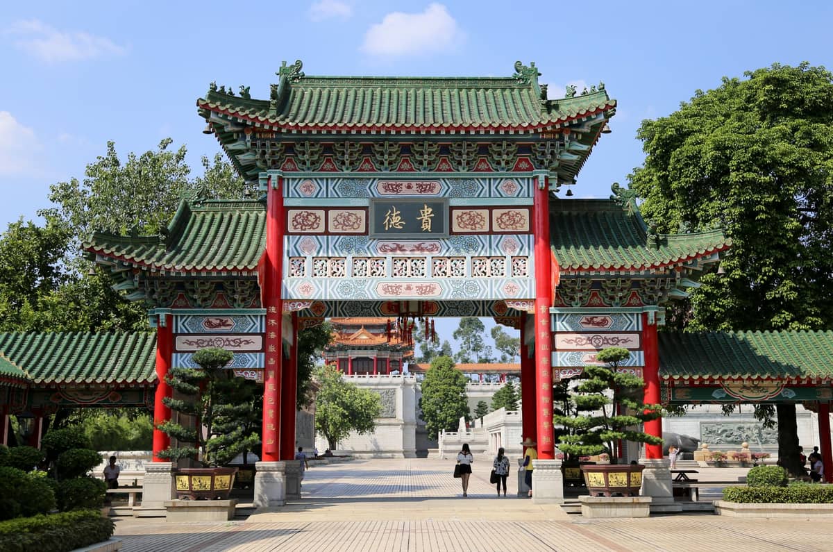 Bright red and green traditional Chinese temple gate with intricate carvings and a pagoda visible in the background, trees on both sides.