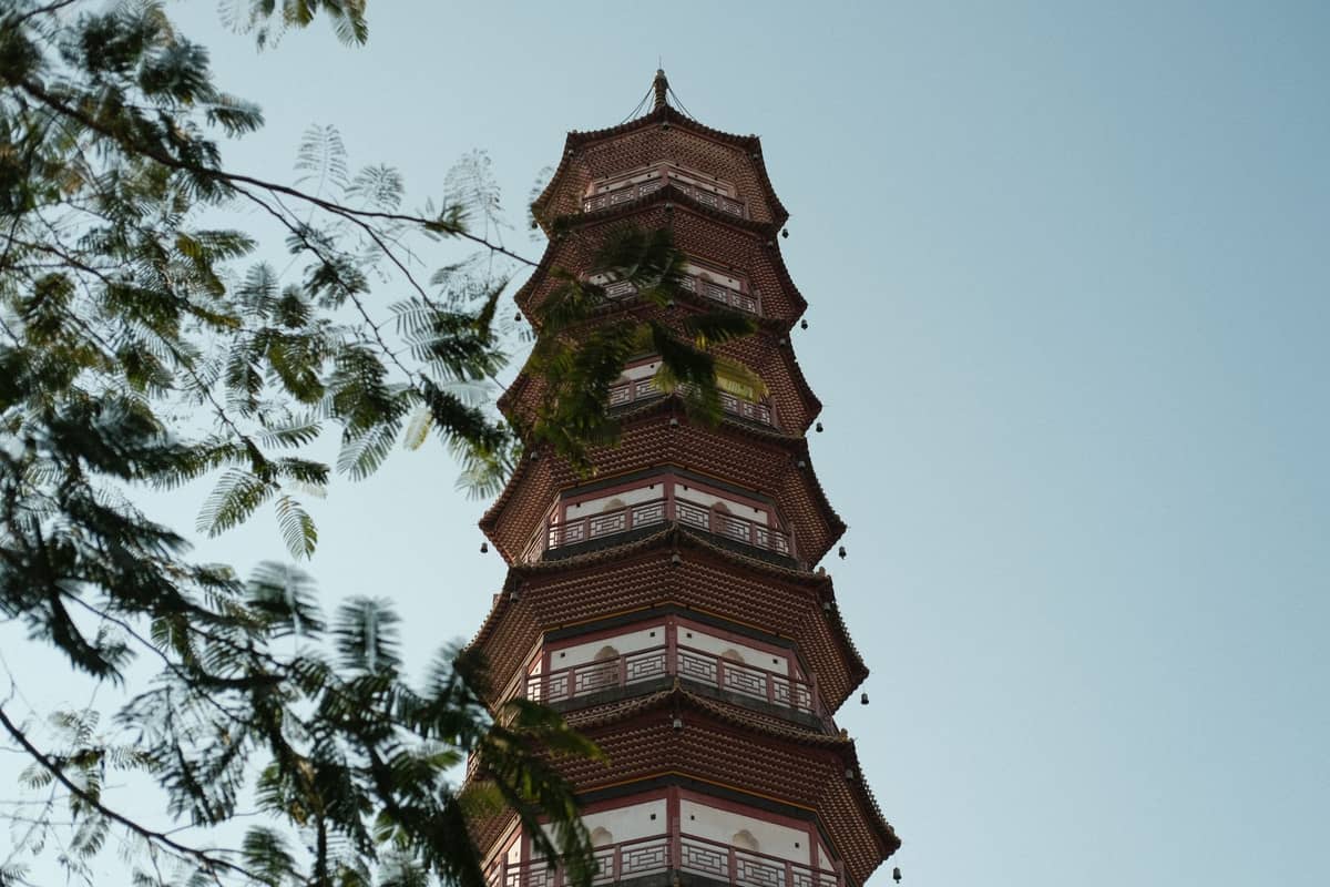 Tall Chinese pagoda with red accents seen from a low angle against a clear blue sky, leafy branches framing the scene.