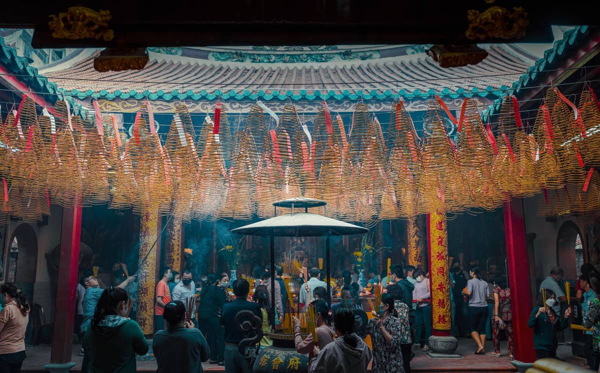Worshippers gather inside a traditional Chinese temple courtyard, filled with incense smoke and spiral incense coils hanging from the roof.