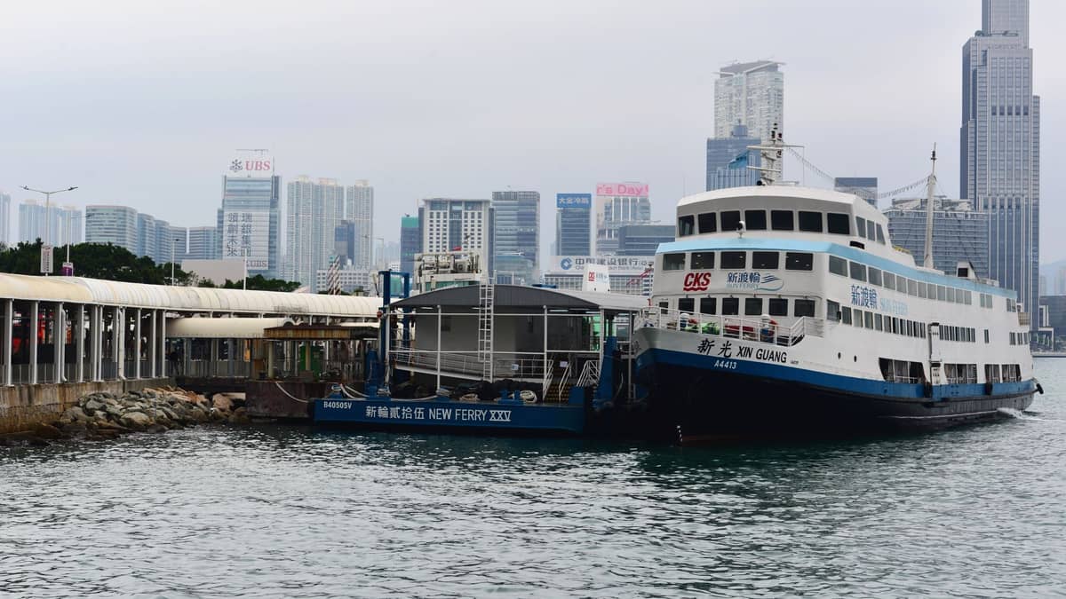 Passenger ferry docked at a terminal with Hong Kong skyline and high-rise buildings in the background.