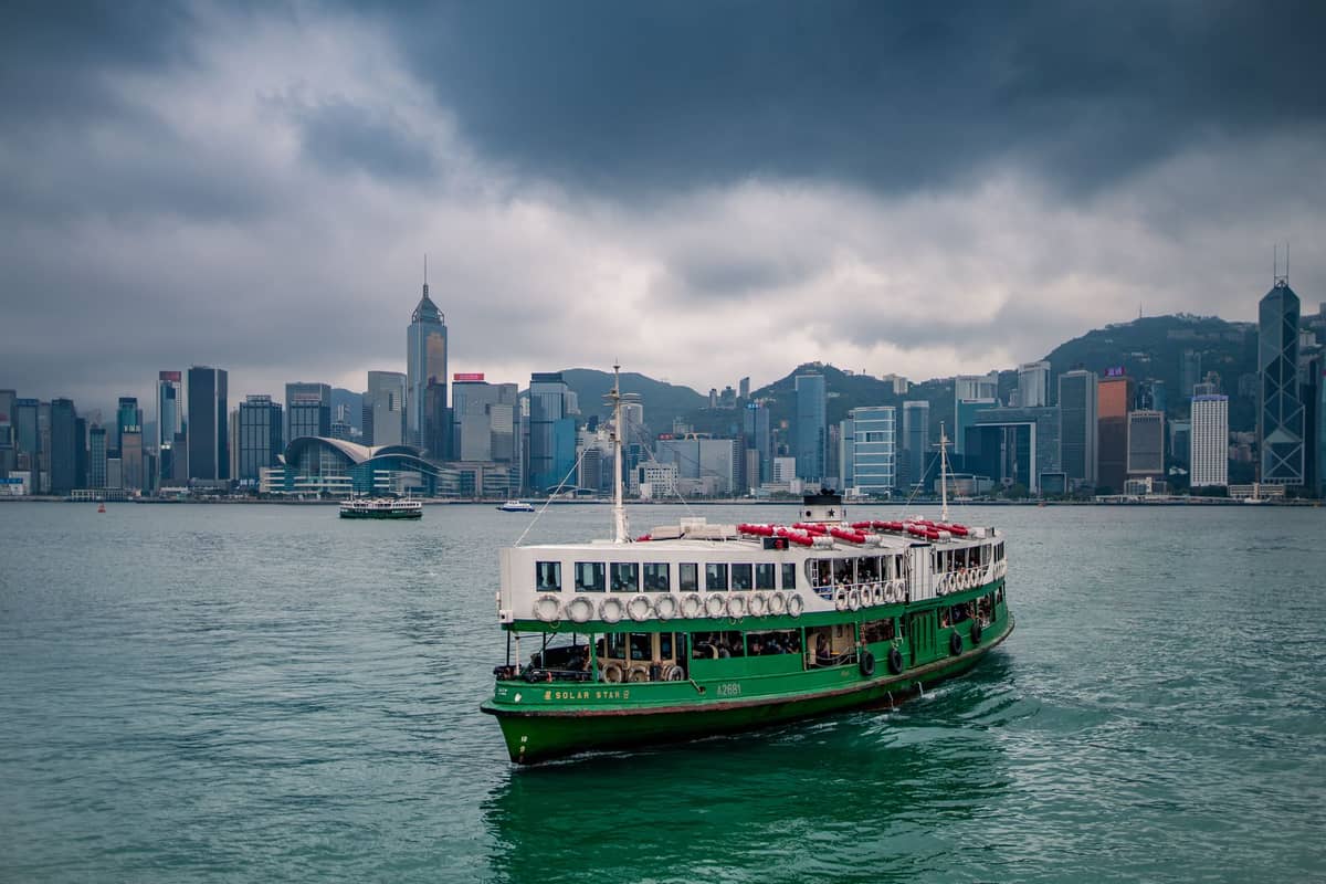 Star Ferry boat crossing Victoria Harbour with the Hong Kong skyline in the background under dramatic cloudy skies.
