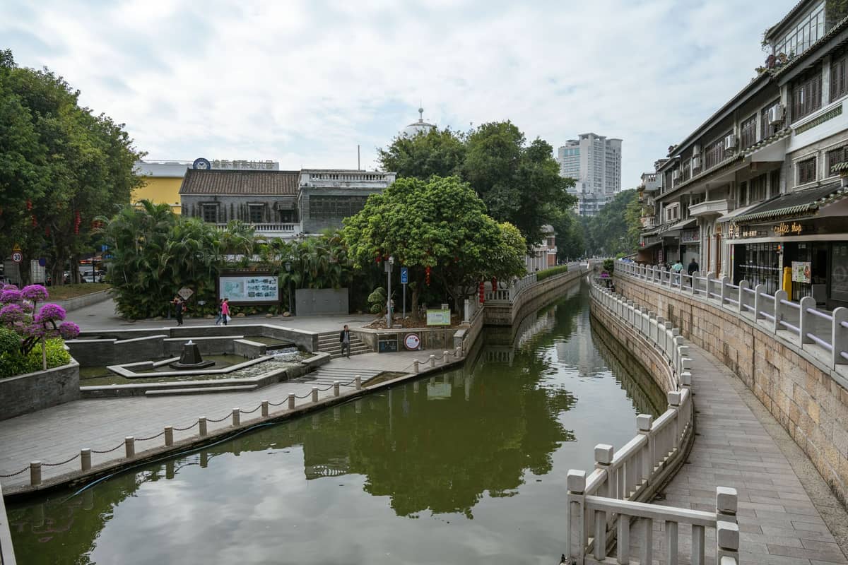 A pedestrian-friendly canal-side walkway in Guangzhou with traditional buildings, greenery, and people walking along the paved path on a cloudy day.