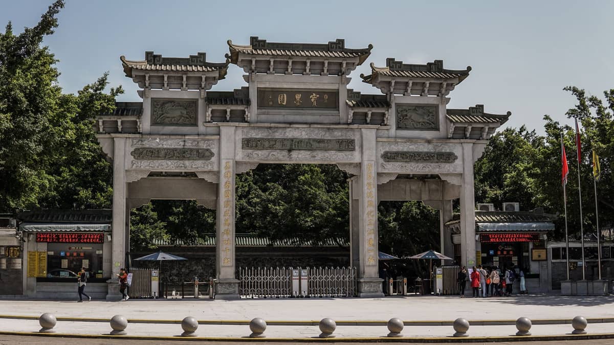 Stone entry gate with traditional Chinese architectural details and characters, people walking nearby, trees and ticket booths in background.