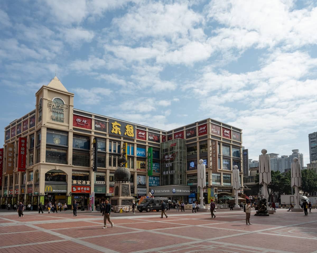 Wide view of a large wholesale market building in Guangzhou with multiple shopfronts and signs in Chinese, people walking in a busy plaza in front.