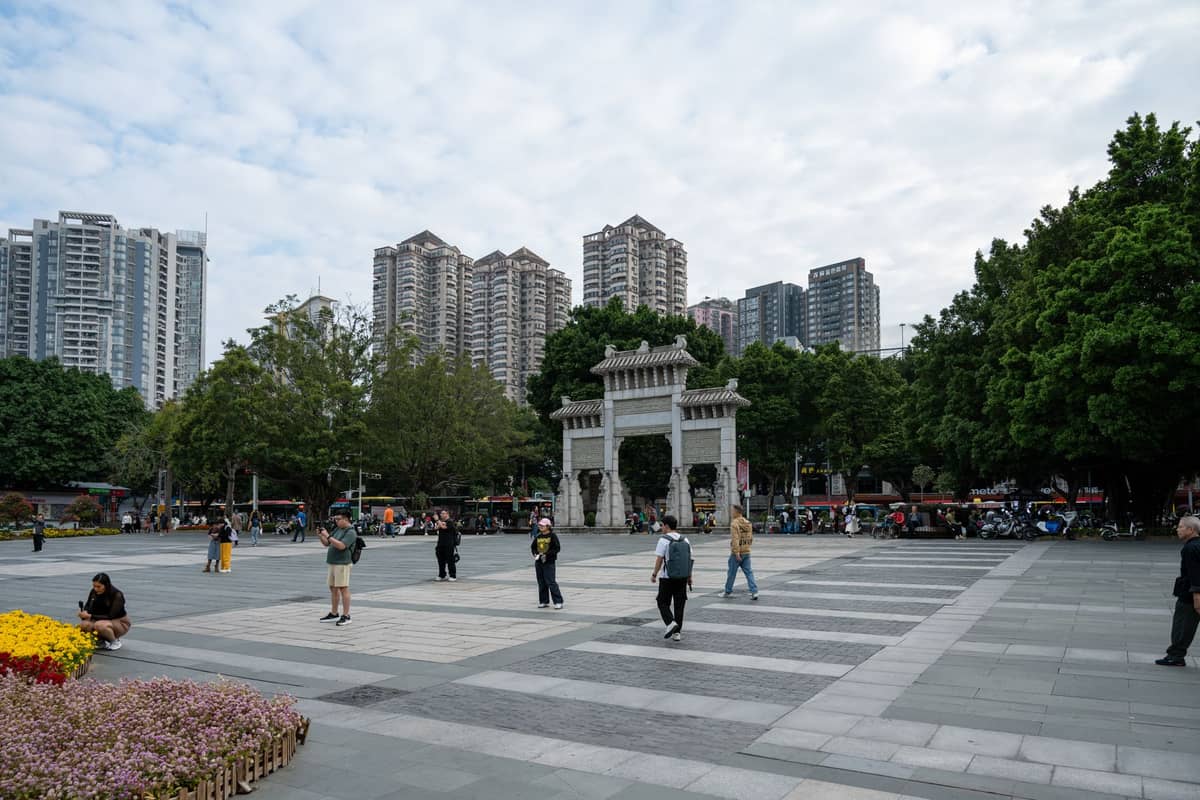 A city plaza in Guangzhou with people walking, a decorative arch, trees, and tall modern apartment buildings in the background.