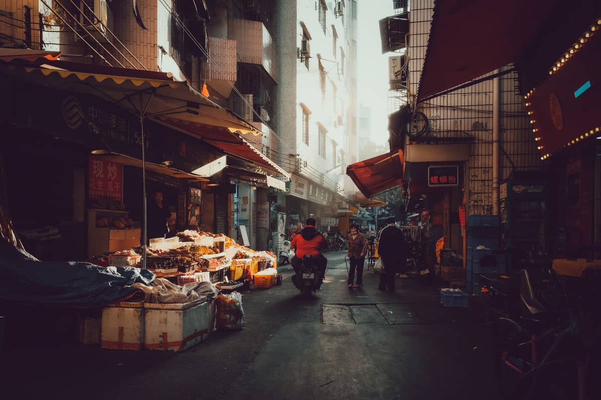View of a bustling narrow market street in Guangzhou, with locals, market stalls, and sunlight filtering between residential buildings.