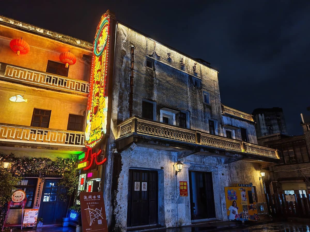 Edifícios históricos de estilo hakka na Cidade Antiga de Guanlan iluminados por letreiros de néon e lanternas à noite, com um céu dramático ao fundo.