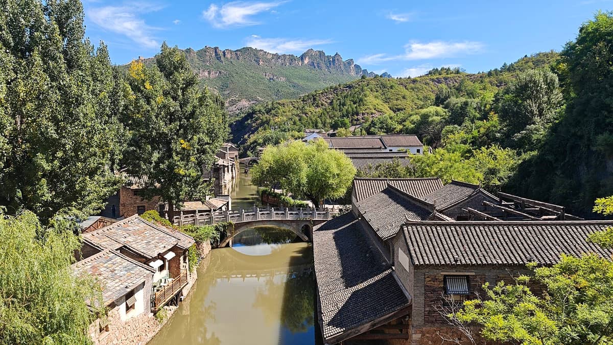 Gubei Water Town mit traditionellen Steinhäusern und Ziegeldächern an einem Kanal, bewaldeten Hügeln und der Simatai-Großen Mauer im Hintergrund unter blauem Himmel.