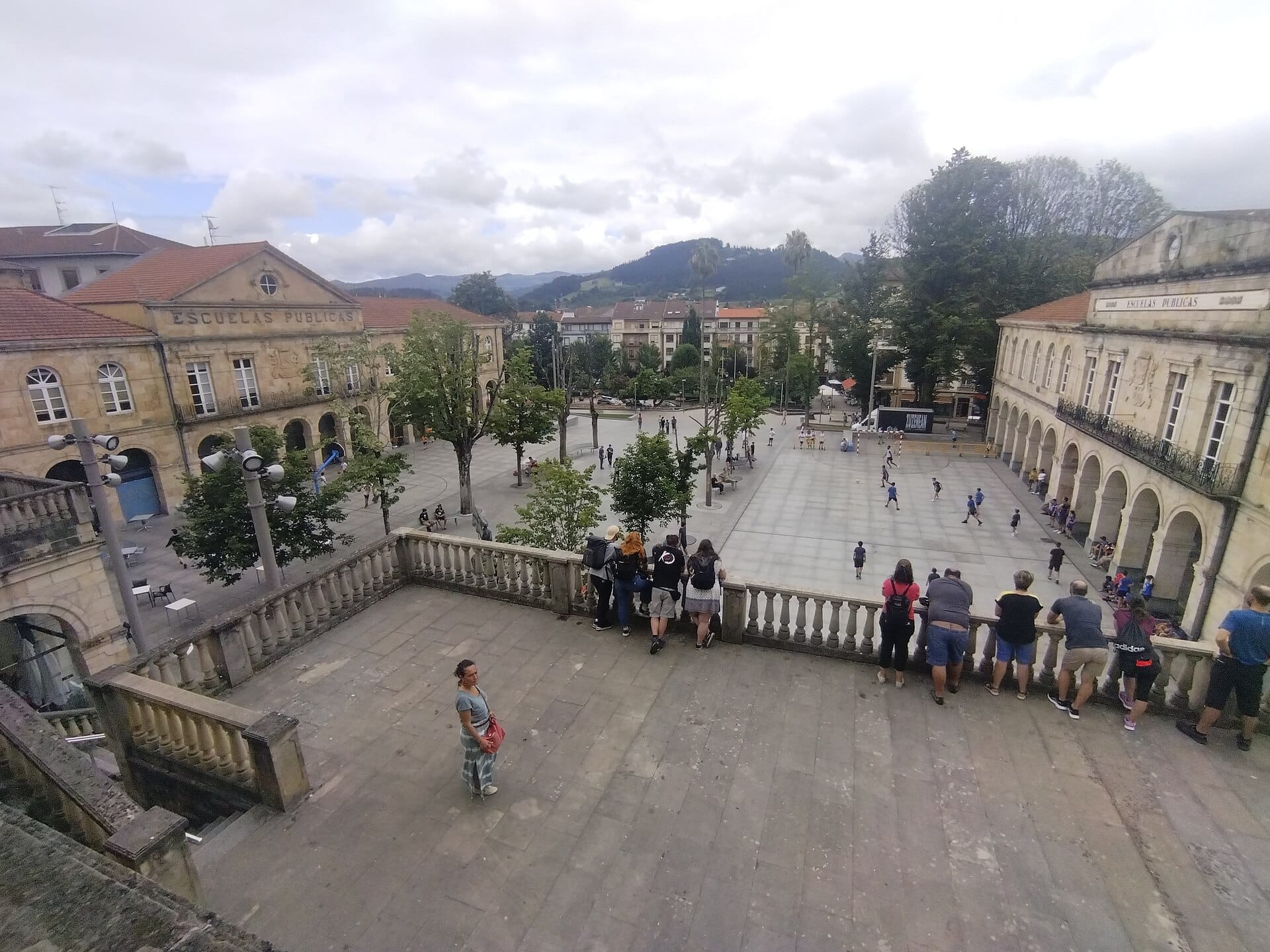 View of Gernika-Lumo’s central square with historic public school buildings, people walking, and distant Basque hills under a cloudy sky.